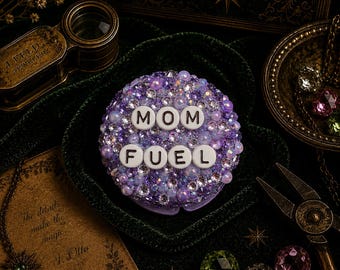Op de afbeelding: Een ronde compacte spiegel versierd met een dichte opstelling van paarse, lavendelkleurige en heldere iriserende strass-steentjes en kralen. Het midden bevat witte ronde kralen met zwarte letters die de tekst MOM FUEL vormen.