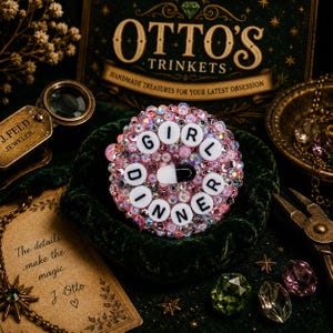 Puede incluir: Un broche rosa e iridiscente con las palabras "GIRL DINNER" escritas con cuentas de letras blancas. El broche está centrado en una superficie de terciopelo verde oscuro, rodeado de joyas y el texto "OTTO'S TRINKETS".