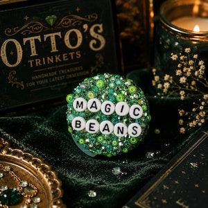 Op de afbeelding: Een ronde, groene snuisterij met de woorden "MAGIC BEANS" in witte letters. De snuisterij is bedekt met groene kralen en edelstenen. De achtergrond omvat een donkergroen fluwelen oppervlak en een decoratieve doos met "OTTO'S TRINKETS".