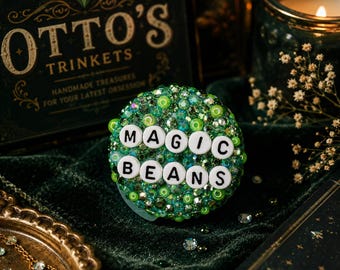 Op de afbeelding: Een ronde, groene snuisterij met de woorden "MAGIC BEANS" in witte letters. De snuisterij is bedekt met groene kralen en edelstenen. De achtergrond omvat een donkergroen fluwelen oppervlak en een decoratieve doos met "OTTO'S TRINKETS".