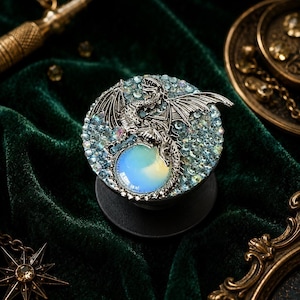 May include: A circular phone grip featuring a silver-toned dragon figure perched atop a large, iridescent, milky-white cabochon stone. The background of the circular face is densely covered in small, light blue, sparkling crystals.