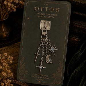 May include: A silver-toned metal charm attached to a dark green display card with the text OTTO'S CHARMS FOR YOUR NEXT CHAPTER and KINDLE CHARM designed with story in mind. The charm features a sword, a star, and a crescent moon with blue stones.