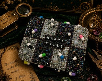 Op de afbeelding: Een rechthoekige clutch bedekt met donkere en lichte glinsterende kristallen in een dambordpatroon. Het oppervlak is versierd met kleine, kleurrijke pilvormige kralen en grotere iriserende parels. De tekst HAPPY PILLS is in het midden gespeld met zwarte letterkralen.