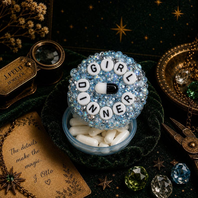 Op de afbeelding: Een ronde, doorzichtige plastic container gevuld met witte capsules. Het deksel is bedekt met lichtblauwe en zilveren strass-steentjes, met witte letterkralen die in een cirkel "GIRL DINNER" spellen, met een zwart-witte capsule in het midden.