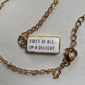 May include: A gold-toned metal chain necklace featuring a rectangular white pendant with a gold-coloured border. The pendant displays the dark blue text "FIRST OF ALL... I'M A DELIGHT" in a simple, sans-serif font. The necklace includes a lobster clasp.