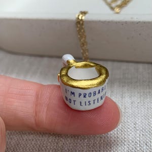 May include: A miniature white ceramic mug pendant on a gold-toned chain. The mug features a metallic gold rim and the black text I'M PROBABLY NOT LISTENING printed on the side. The pendant is suspended from a delicate gold-coloured metal link chain.