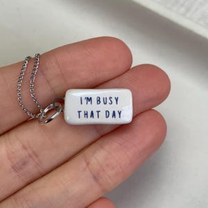 May include: A rectangular white ceramic pendant on a silver-toned metal chain. The pendant features the dark blue text "I'M BUSY THAT DAY" printed on its front surface. The piece is attached to the chain with a small silver-toned metal jump ring.