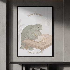 Può includere: Una stampa artistica incorniciata raffigurante una tartaruga verde seduta a una scrivania di legno, intenta a scrivere su un libro aperto con una candela accesa accanto. Il testo "Write it Down" appare sopra la tartaruga e "And Let it Go" sotto.