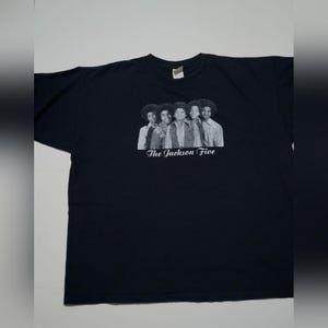 May include: A black short-sleeved t-shirt featuring a centered black and white photographic print of the musical group The Jackson Five. Below the image, the text The Jackson Five is printed in a white script font on the front of the shirt.