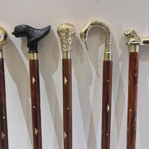 May include: Five walking sticks with dark brown wooden shafts featuring inlaid brass accents. The handles include a polished brass sphere, a black animal head, an ornate brass knob, a textured brass swan neck, and a polished brass lion figure.