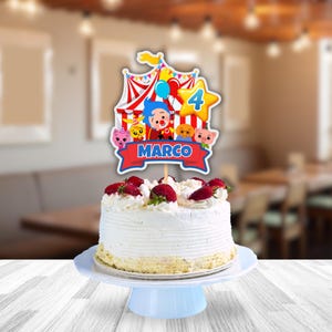 Puede incluir: Una tarta de cumplea&ntilde;os con glaseado blanco, cubierta con fresas frescas. Un adorno de tarta con tem&aacute;tica de circo colorido con personajes de dibujos animados, el n&uacute;mero 4 y el nombre "MARCO" en una pancarta roja. La tarta est&aacute; sobre un soporte azul claro.