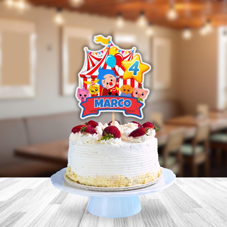 Puede incluir: Una tarta de cumplea&ntilde;os con glaseado blanco, cubierta con fresas frescas. Un adorno de tarta con tem&aacute;tica de circo colorido con personajes de dibujos animados, el n&uacute;mero 4 y el nombre "MARCO" en una pancarta roja. La tarta est&aacute; sobre un soporte azul claro.