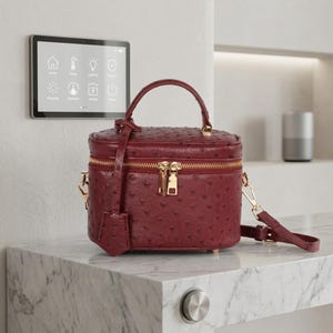 May include: A burgundy ostrich-textured vanity case with a gold zipper and hardware. The small handbag has a top handle and a detachable shoulder strap. A small tag hangs from the zipper. The bag sits on a marble surface.