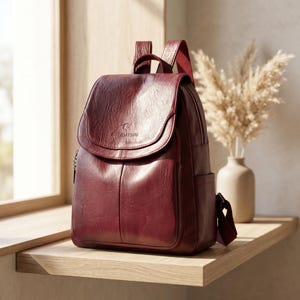May include: A burgundy leather backpack with a flap closure and a top handle. The backpack has a front pocket and side pocket. The brand name "BALIDATSHU" is visible on the flap. The backpack is sitting on a wooden shelf.
