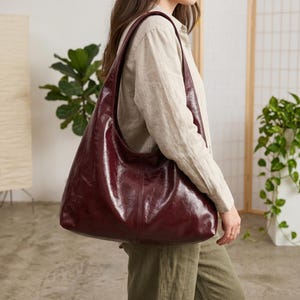 May include: A large, burgundy leather shoulder bag with a curved design. The bag has a single shoulder strap and a glossy finish. The bag is worn over the shoulder. The bag is a deep red color.