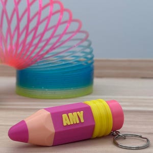 Könnte beinhalten: Ein bleistiftförmiger Schlüsselanhänger mit einer rosa Spitze, einem pfirsichfarbenen Abschnitt, einem magentafarbenen Körper mit dem Namen AMY in gelben Buchstaben und einem gelben gerippten Ende. Dahinter liegt eine bunte Spiralfeder.