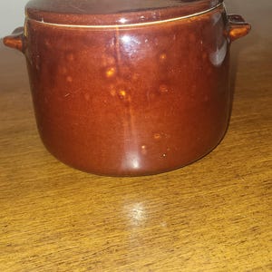 May include: A round, dark brown glazed ceramic pot with a matching lid and two small side handles. The exterior surface features a glossy finish with subtle, lighter brown mottled spots throughout the glaze, creating a textured appearance.