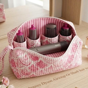 Puede incluir: Un estuche de almacenamiento acolchado con estampado rosa y blanco y cierre de cremallera, que contiene una herramienta de peinado Dyson gris y cuatro accesorios cilíndricos. El interior tiene un forro a rayas rosas y blancas.