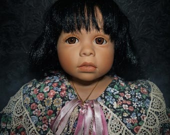 May include: A porcelain doll with black hair and brown eyes. The doll wears a floral dress with a lace collar and a pink ribbon necklace. The background is a dark, patterned fabric.