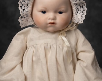 May include: A porcelain doll with blue eyes, wearing a cream-colored dress and a matching bonnet with lace trim. The doll has rosy cheeks and a serious expression, set against a dark gray backdrop.