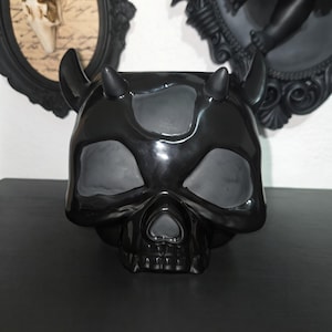 May include: A glossy black ceramic skull planter featuring two small horns on the top. The skull has hollowed-out eye sockets, a nasal cavity, and a top opening, all finished in a smooth, reflective black glaze that highlights its distinct shape.