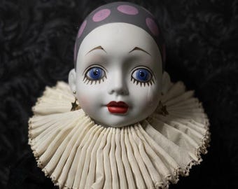 May include: A porcelain doll head with a gray cap featuring pink polka dots. The doll has blue eyes, red lips, and a cream-colored ruffled collar. Gold star earrings complete the look. The doll is dressed in a black outfit.