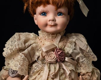 May include: A porcelain doll with red hair styled in two buns and blue eyes. The doll wears a cream-colored dress with lace detailing, ribbons, and rose accents. The doll has a serene expression and is set against a black background.