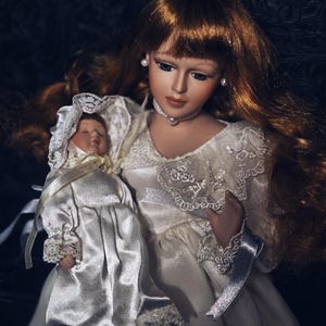 May include: A porcelain doll with long auburn hair and a white satin dress, holding a smaller doll in a matching outfit. The larger doll wears a pearl necklace and earrings. The dolls are set against a dark, textured background.