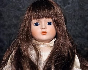May include: A porcelain doll with long brown hair and blue eyes. The doll has a fair complexion, rosy cheeks, and a small, orange-red mouth. She is wearing a white dress with a lace collar.