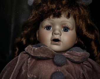May include: A vintage porcelain doll with blue eyes and auburn hair. The doll wears a mauve velvet dress with a dark gray collar and two matching buttons. A large gray bow adorns the doll's head.
