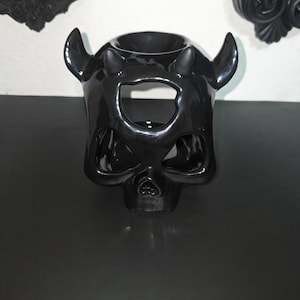 May include: A glossy black ceramic wax warmer shaped like a stylized skull with two prominent horns on top. The design features cutouts for the eyes and nose, with a circular basin at the crown for holding wax melts or essential oils.