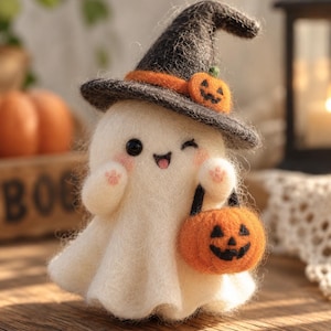 Pode incluir: Uma figura de fantasma branco em feltro com um chapéu de bruxa preto, uma faixa laranja e um pequeno detalhe de abóbora. O fantasma tem bochechas rosadas, um olho a piscar, um olho preto e segura um pequeno balde de abóbora em feltro.