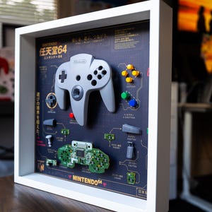 May include: A white shadow box frame containing a disassembled grey Nintendo 64 controller. The display features the controller shell, internal green circuit board, and various buttons arranged on a dark background with Japanese text and technical diagrams.