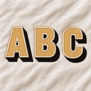 Puede incluir: Tres letras bordadas A, B y C en hilo de color dorado con un borde blanco grueso y un efecto de sombra paralela negra. Las letras están cosidas sobre un fondo de tela texturizada de color blanco roto, creando una apariencia dimensional.