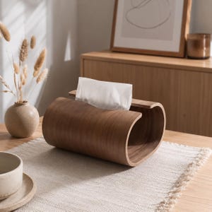 Puede incluir: Un soporte para pañuelos de madera de color marrón oscuro curvado con un acabado liso y pulido. El soporte rectangular presenta un diseño de extremos abiertos que se curva hacia adentro en los lados, con un pañuelo blanco que sobresale.