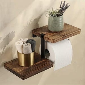 Peut inclure: Une étagère murale composée de deux tablettes en bois foncé reliées par un cadre en métal noir. La tablette supérieure supporte un pot en céramique vert clair moucheté avec des plantes séchées. La tablette inférieure contient un gobelet en métal doré.
