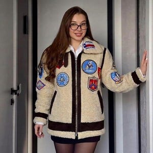 Puede incluir: Una chaqueta de cremallera color crema, de textura peluda, con ribetes marrón oscuro y varios parches bordados, incluyendo una bandera de Texas con la inscripción 'DALLAS' en el cuello y un parche circular 'SEABEES' en el pecho.