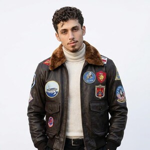 May include: A dark brown leather bomber jacket featuring a plush brown fur collar, a front zipper closure, and two flap pockets. The jacket is adorned with various colorful embroidered military-style patches on the chest and sleeves.
