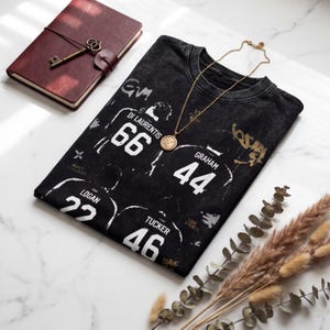 Könnte beinhalten: Schwarzes T-Shirt mit weißen Spielernummern und -namen, darunter "Di Laurentis 66" und "Graham 44." Eine goldene Anhängerkette liegt auf dem Shirt. Ein weinrotes Lederjournal mit Schlüssel und Trockenblumen sind ebenfalls zu sehen.
