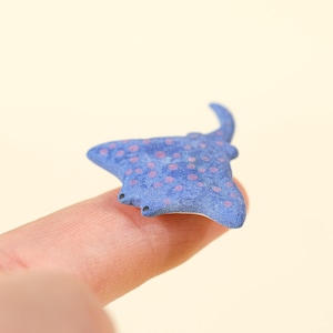 Peut inclure: Une petite figurine de raie bleue, plate, présentant une surface texturée avec des pois roses dispersés. La raie possède deux petits yeux noirs situés sur le bord avant et une queue courbée s'étendant à l'arrière de son corps triangulaire.
