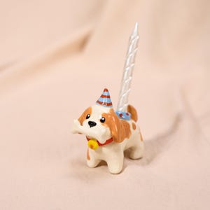 Könnte beinhalten: Eine kleine Keramik-Hundefigur mit braunen und weißen Flecken, die ein rotes Halsband mit einer gelben Glocke und einen blau-rot gestreiften Partyhut trägt. Der Hund hält einen weißen Knochen im Maul und hat eine hohe, gedrehte weiße Kerze am Rücken.