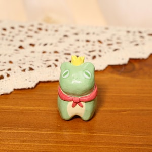 May include: A small, green frog figurine with a yellow crown and a red bow. The frog has oval eyes and a light green body. The figurine is sitting on a wooden surface.