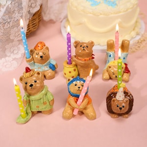 May include: A collection of six ceramic bear figurines, each holding a lit birthday candle with white polka dots. The bears are dressed in various outfits, including hats and scarves, and feature painted details like flowers and patterns on their clothing.