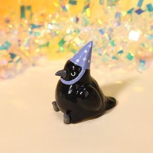 Puede incluir: Una pequeña figura de pájaro de cerámica negra brillante con un cuerpo liso y redondeado y un pico puntiagudo. El pájaro lleva un sombrero de fiesta de color morado claro decorado con pequeñas estrellas blancas y tiene dos pies grises.