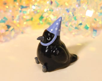 Op de afbeelding: Een klein, glanzend zwart keramieken vogelbeeldje met een glad, rond lichaam en een spitse snavel. De vogel draagt een lichtpaarse feesthoed versierd met kleine witte sterpatronen en heeft twee kleine grijze voetjes aan de onderkant.