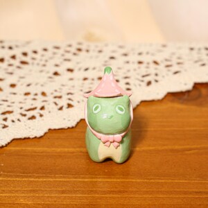 Pode incluir: Uma figura de sapo de cerâmica pintada à mão. O sapo é principalmente verde com um chapéu e cachecol rosa. A figura tem olhos grandes e ovais e uma pequena fita. O sapo está sobre uma superfície de madeira.
