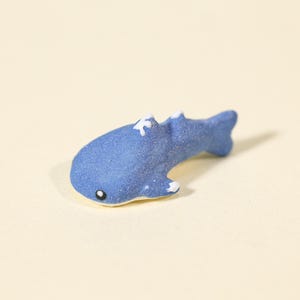 May include: A small, textured blue whale figurine with a matte finish. The whale features a rounded head, a single black eye with a white pupil, and small white accents on the dorsal fin and pectoral fins. The underside of the whale is cream-colored.