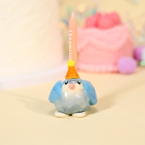 May include: A light blue, round, bird-shaped candle holder featuring a small orange and yellow striped party hat on its head. A tall, thin, pale pink birthday candle is inserted into the top of the bird, with the word "birthday!" printed in white.