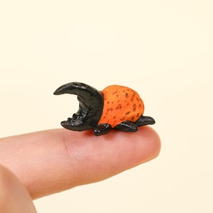 May include: A miniature beetle figurine featuring a glossy black head and thorax with a prominent horn, paired with a textured, bright orange abdomen marked by small dark brown spots, resting on a fingertip to show its small scale.
