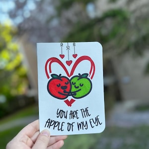 May include: A white greeting card featuring a red and a green apple character hugging inside a red heart outline. Above the apples are three small red hearts hanging from strings. The text reads, "YOU ARE THE APPLE OF MY EYE" in black.